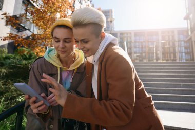 İki kadın, taş bir merdivenin ve büyük bir binanın zeminine karşı telefonda zevkle bakıyorlar.