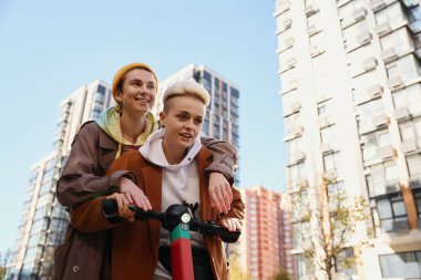Neşeli genç kadınlar, lgbt çiftleri, sonbahar güneşli bir günde şehirde scooter 'la geziyorlar, gökdelenlerin arka planına karşı.