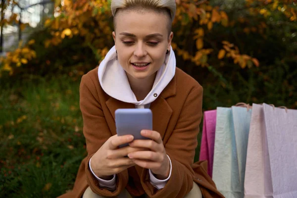 코트를 금발의 여성은 공원에서 휴식을 취하고 그녀는 전화로 메시지를 — 스톡 사진