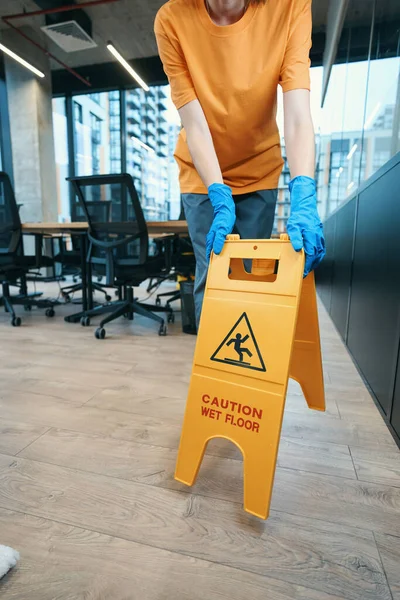 Cleaner in overalls and rubber gloves puts a yellow wet floor warning sign in an office area