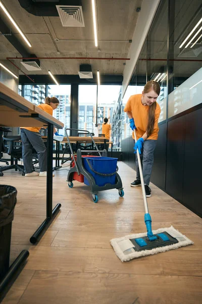 Team of cleaners in overalls and rubber gloves work in a coworking area, women use a mop, buckets, rags