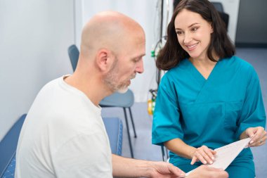 Tıbbi üniformalı neşeli bir kadın, elindeki çıktıya dikkatlice bakarken yanında oturan adama bakar.