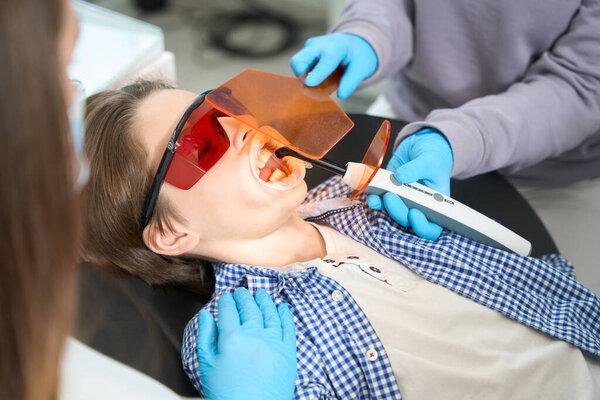 Young patient in protective glasses lies in a dental chair, dentist with an assistant seals a tooth to a teenager