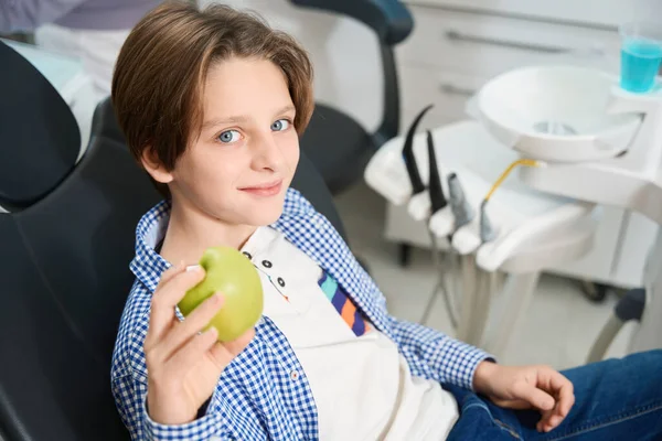 笑着的男孩坐在牙医椅上 拿着特殊的工具 手里拿着一个苹果 — 图库照片