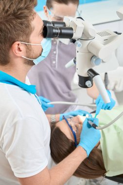 Hemşire eşliğinde mikroskop altında dişlerini tedavi eden deneyimli bir erkek doktor.