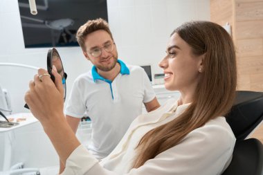 Gülümseyen dişçi kliniği müşterisi neşeli bir dişçinin huzurunda ameliyat sonrası aynaya bakıyor.