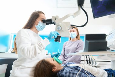 Modern bir diş kliniğinde kök kanal tedavisi anestezi ile yapılır, mikroskop altında, dişçi asistanıyla çalışır.