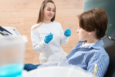 Kadın dişçi genç bir hastayla kibarca iletişim kurar. Elinde özel aletler vardır.