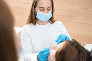 İşyerindeki dişçi kadın bir çocuğa yardım ediyor, bir asistanla çalışıyor.