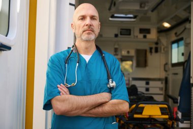 Mavi üniformalı katı bir doktor, kollarını göğsüne kavuşturmuş ve arkasında bir tıp uzmanının kamarası duruyor.