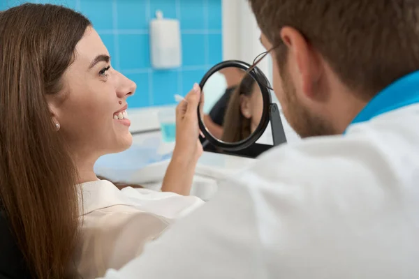 愉快的女病人手拿着镜子坐在椅子上看牙医 — 图库照片