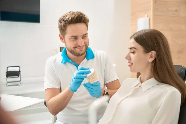 Leende Vänlig Tandläkare Visar Typodont Till Ung Kvinnlig Patient Sittande — Stockfoto