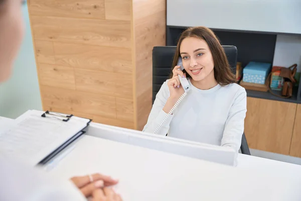 stock image Smiling friendly secretary seated at reception desk looking at client during phone conversation