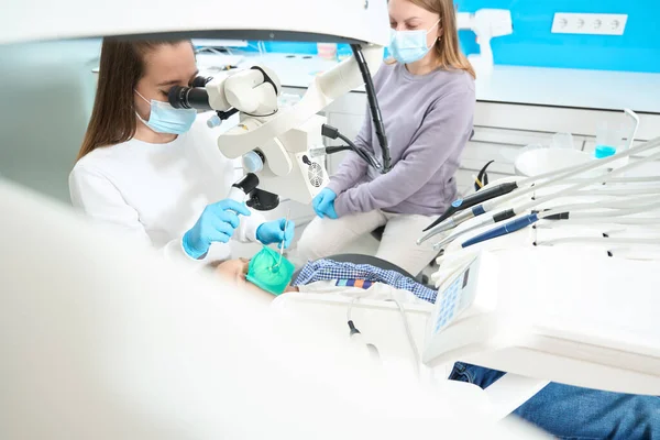 Root canal treatment for child in modern dental clinic is carried out with anesthesia, under microscope, dentist works with assistant