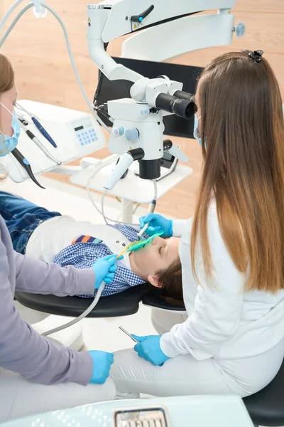 女牙医和一名助手正在用现代设备为男孩修理一颗牙 并使用显微镜 — 图库照片