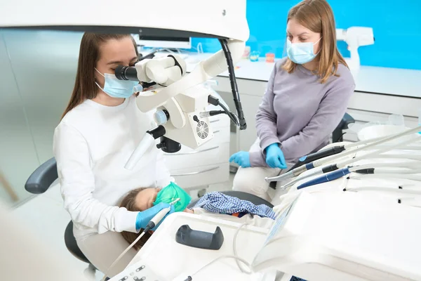 牙医正在显微镜下治疗一个男婴牙 她用的是现代设备 旁边有一个助手 — 图库照片