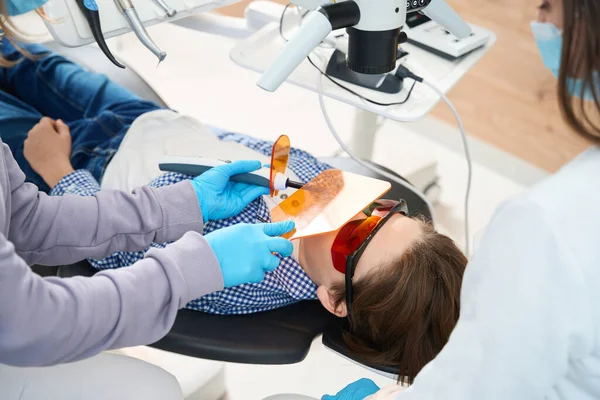 戴防护眼镜的孩子躺在牙医的椅子上 医生和助手给他填饱了肚子 — 图库照片