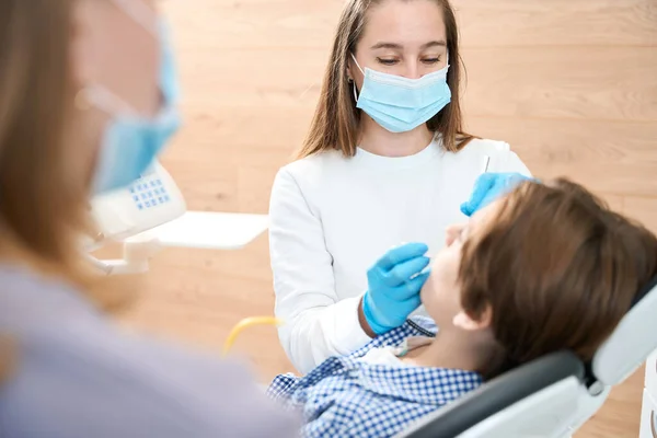 牙医妇女正在一家牙科诊所协助一个男孩 她正在与一名助手一起工作 — 图库照片