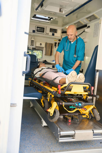 Serious man fastening seat belts in ambulance while patient lies on her back with arms outstretched along body