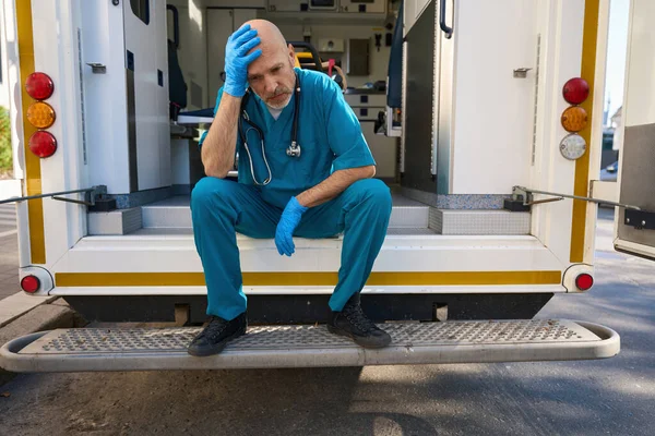 現代の救急車の階段に座っている間に悲しみを経験する医療の制服を着た疲れた男 — ストック写真