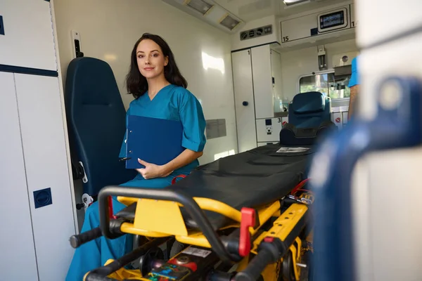 満足した看護師は 彼女の手にクリップボードを保持しながら 救急医療のための車のサロンのガーニーに座っています — ストック写真