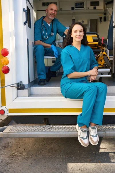 身穿医疗制服的心满意足的女人坐在一辆救护车上的担架旁边 而一个戴着不孕手套的男人蹲在旁边 — 图库照片