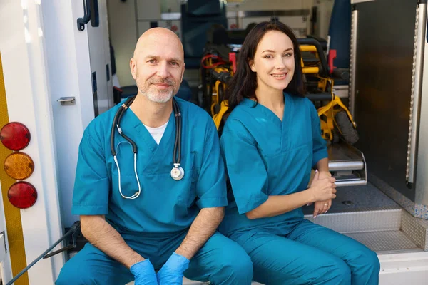 一位快乐的救护车妇女坐在医疗运输的边缘上 而她的同事则手拿无菌手套坐在她旁边 — 图库照片