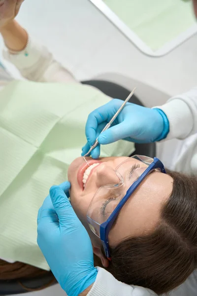 歯医者が彼女の前の歯を検査しながら 女性患者は ビブと保護アイウェアを着て — ストック写真