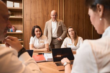 Klinik müdürü ofiste, masada bir toplantı düzenliyor. Bir dizüstü bilgisayar, belgeler, dosyalar.