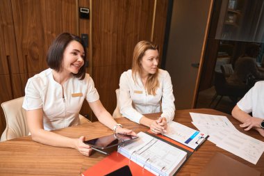 Kadın doktor genel bir toplantıda iş arkadaşlarıyla iletişim kuruyor, kızıl saçlı bir kadın da yanında oturuyor.