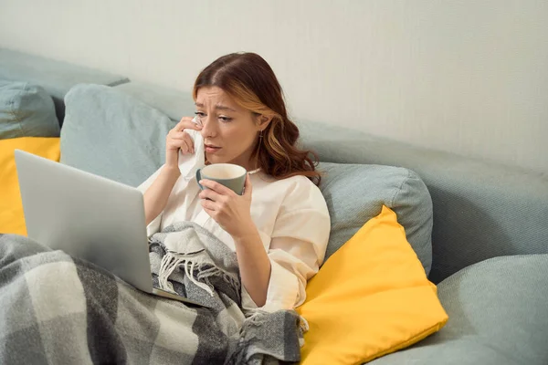 Upset young woman seated in bed in front of laptop wiping away tears with handkerchief
