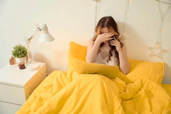 Woman seated with laptop in bed covering her eyes with hand during phone conversation