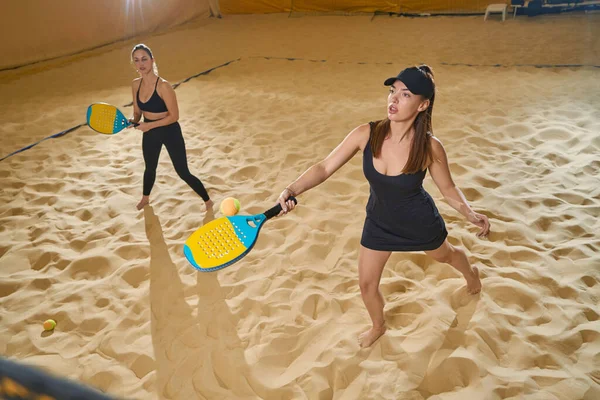 穿着运动服的严肃女性赤脚站在沙滩上 手里拿着球拍 旁边有一个玩伴在看着 — 图库照片