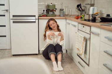 Smiling pleased woman sitting alone on kitchen floor with ceramic cup in hands