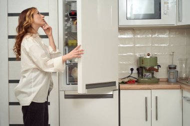 Side view of calm focused young female standing before open fridge door and looking inside