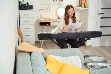Concentrated female composer playing synthesizer while staring at sheet music in hand