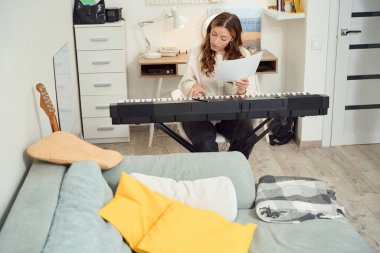 Focused female keyboardist holding sheet music in her hand and playing synth