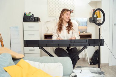 Smiling cheerful young music teacher performing musical composition on synthesizer before smartphone camera