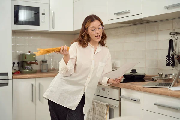 集中女性は台所のカウンターで肘を傾け手にスパゲティを保持し 紙のシートを見て — ストック写真
