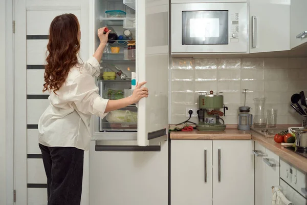 台所に立つ女性のバックビューと冷蔵庫の棚にドリンクとボトルのために達する — ストック写真