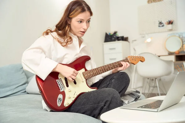Focused woman seated on bed brushing fingers over strings of guitar while looking at laptop screen