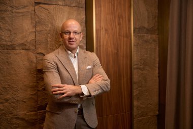 Bald, benevolent middle-aged man stands against the wall, arms crossed over his chest, he is in a beautiful room