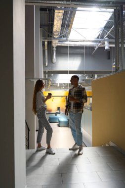 Full-length photo of man and woman talking while standing in the office, holding coffee