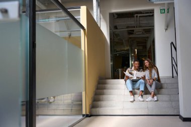 Smiling coworkers sitting on steps and examining the documents in the office