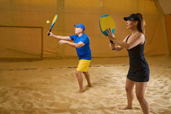 注目すべき女性でスポーツウェアで彼女のパートナーを見ていますビーチテニスをしながら彼はテニスボールを打つ — ストック写真