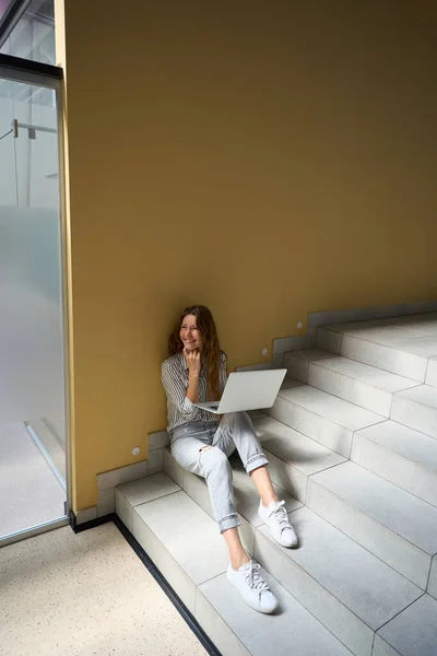 Top view of happy lady working with laptop while sitting on steps indoors