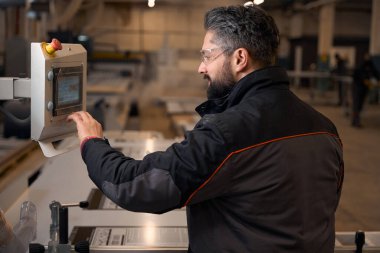 Male in protective clothes and glasses standing near computer of form cutting machine, selecting operating program