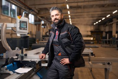 Happy man in protective clothes and glasses standing near computer in workshop, looking at the camera