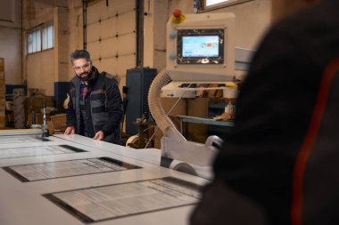 Professional worker in protective clothes and glasses standing near form cutting machine and cutting material