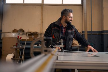 Male in protective clothes and glasses standing near woodworking machine and cutting material in workshop
